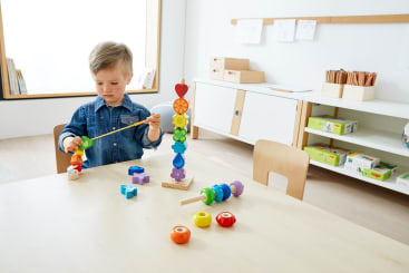 Ein kleines Kind in einem Jeanshemd sitzt an einem Tisch in einem hellen Klassenzimmer und fädelt bunte Holzperlen auf einen Stock. Auf dem Tisch und in den Regalen im Hintergrund sind verschiedene Lernspielzeuge und Kunstmaterialien zu sehen.