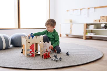 Ein kleiner Junge in einem grünen Hemd sitzt auf einem grauen Teppich und spielt mit einer Spielzeugburg aus Holz und mehreren Ritter- und Drachenfiguren. Der Raum ist mit hellen Möbeln und großen Fenstern ausgestattet.