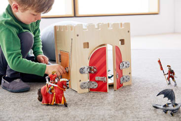 Ein kleines Kind in einem grünen Pullover spielt mit einer Spielzeugburg aus Holz. Spielzeugfiguren von Rittern und ein Drache sind um die Burg herum auf einem Teppichboden angeordnet. Durch große Fenster im Hintergrund fällt Licht ein.