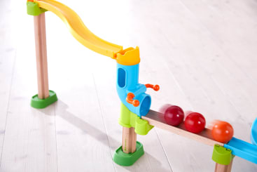 Eine hölzerne Murmelbahn mit einer gelben Bahn, blauen und grünen Stützen und drei farbigen Kugeln - rot, orange und rot - auf einer hölzernen Rampe auf einem hellen Holzboden.