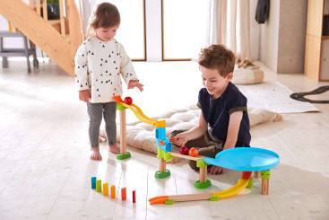 Zwei kleine Kinder spielen mit einer bunten Kugelbahn auf einem hellen Holzboden. Die Bahn verfügt über Rampen, eine Spirale und Dominosteine, die am Ende aufgestellt sind. Im Hintergrund sind Kissen und Holzmöbel zu sehen.