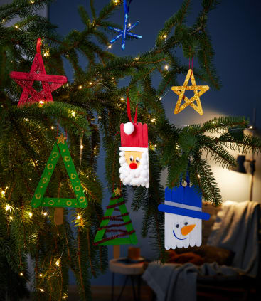 Ein Tannenzweig, geschmückt mit handgefertigtem Weihnachtsschmuck, darunter Sterne, Bäume, ein Weihnachtsmann und ein Schneemann, alles aus bunten Eisstielen, mit Lichterketten im Hintergrund.