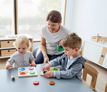 Eine erwachsene Frau beobachtet zwei kleine Kinder, die an einem Tisch sitzen und in einem hellen, aufgeräumten Raum mit Puzzles und bunten Spielfiguren spielen.