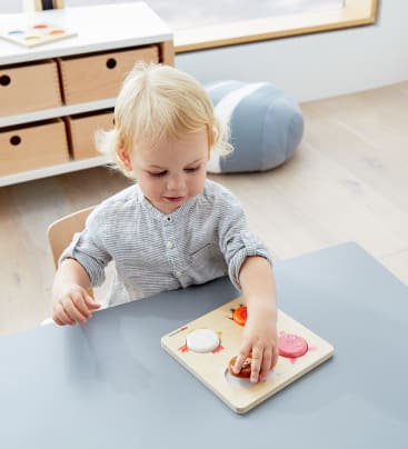 Ein kleines Kind mit blondem Haar sitzt an einem grauen Tisch und legt ein hölzernes Puzzlestück auf ein Puzzlebrett. Im Hintergrund sind ein weiches blaues Kissen und hölzerne Aufbewahrungsschubladen zu sehen.