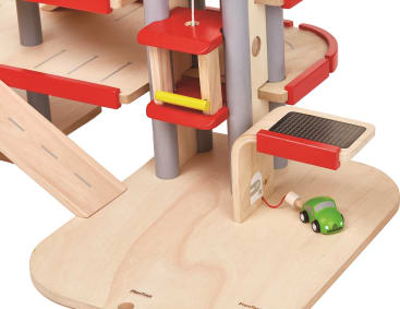 Ein Spielzeugparkhaus aus Holz mit rotem Geländer, grauen Säulen, einem gelben Aufzug und einem grünen Spielzeugauto, das an eine Ladestation angeschlossen ist. Eine Rampe und ein Teil mit einem schwarzen Solarpanel sind ebenfalls zu sehen.