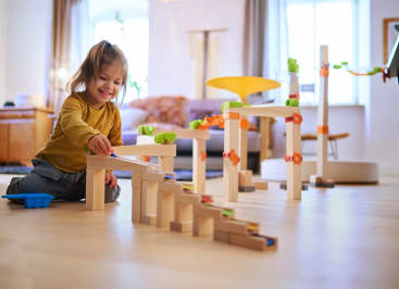 Ein kleines Kind sitzt auf einem Holzfußboden in einem Haus und spielt mit einer Murmelbahn aus Holzklötzen und bunten Plastikteilen. Das Sonnenlicht fällt durch große Fenster im Hintergrund.