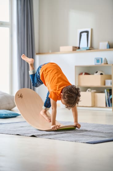 Ein Kind in orangefarbenem Hemd und Jeans balanciert auf Händen und einem Fuß auf einem hölzernen Balancebrett in einem hellen Wohnzimmer. Im Hintergrund befinden sich Regale mit Büchern, Körben und Dekorationsgegenständen.