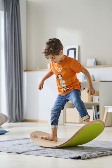 Ein kleiner Junge in orangefarbenem T-Shirt und Jeans balanciert barfuß auf einem geschwungenen Holzbalancierbrett, das auf einem Teppich in einem hellen, modernen Raum steht.