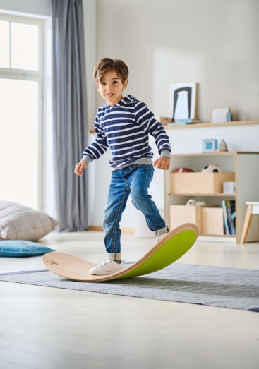 Ein kleiner Junge in gestreiftem Hemd und Jeans balanciert auf einem geschwungenen Holzbalancierbrett in einem hellen, modernen Raum mit Regalen, Kissen und großen Fenstern im Hintergrund.