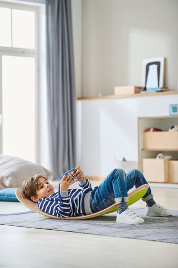 Ein Junge in gestreiftem Hemd und Jeans liegt auf einem gebogenen Balance Board in einem Wohnzimmer und hält einen Videospiel-Controller. Der Raum hat helle Wände, ein Fenster mit Vorhängen und Regale mit Kisten und Dekoration.