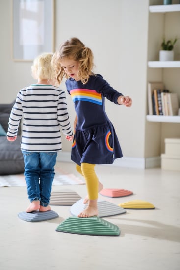 Zwei kleine Kinder balancieren barfuß auf bunten, strukturierten Trittsteinen in einem hellen, aufgeräumten Wohnzimmer. Ein Kind trägt gelbe Leggings und ein marineblaues Kleid mit Regenbogenstreifen, das andere trägt Jeans und ein gestreiftes Hemd.
