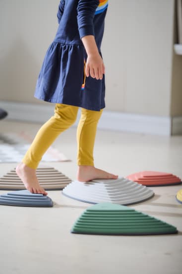 Ein Kind, das ein blaues Kleid und gelbe Leggings trägt, läuft barfuß auf strukturierten, bunten Trittsteinen, die auf einem hellen Boden angeordnet sind.