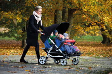 Eine Frau mit dunklem Mantel und Schal schiebt einen großen Kinderwagen mit drei kleinen Kindern, die nebeneinander sitzen. Im Hintergrund sind Bäume mit Herbstlaub und ein mit Laub bedeckter Boden zu sehen.