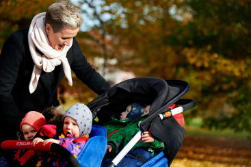 Ein Erwachsener steht im Freien neben einem Kinderwagen mit vier kleinen Kindern, die alle in warme Kleidung gekleidet sind. Der Hintergrund zeigt Herbstlaub mit orangefarbenen und gelben Blättern.