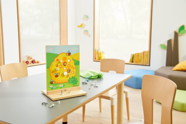 Ein Klassenzimmer mit Holzstühlen und einem Tisch, auf dem ein Brettspiel mit Bienen und Waben abgebildet ist. Im Hintergrund befinden sich Bücherregale, Bücher und bunte Kissen an hohen Fenstern.