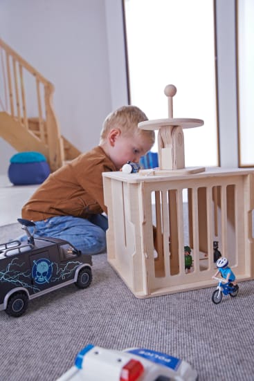 Ein kleines Kind sitzt auf dem Boden und spielt mit einem Spielzeuggefängnis aus Holz und mehreren Spielzeugfahrzeugen, darunter ein Polizeiwagen und ein Motorrad, in einem modernen, gut beleuchteten Raum mit einer Treppe im Hintergrund.