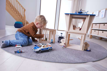 Ein kleiner Junge sitzt auf einem runden grauen Teppich und spielt mit einem Holzspielzeug-Knast-Set, einem Polizeiauto und kleinen Action-Figuren in einem hellen, modernen Raum mit Holzböden und Treppen im Hintergrund.