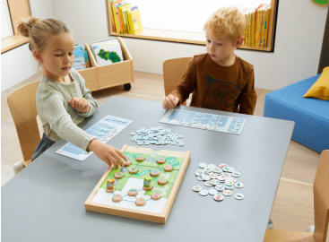Zwei Kinder sitzen an einem Tisch und spielen ein Brettspiel mit bunten, kreisförmigen Figuren. Im Hintergrund befinden sich Regale mit Büchern und ein blauer Sitz. Das Mädchen streckt die Hand aus, um eine Figur auf dem Spielbrett zu bewegen.
