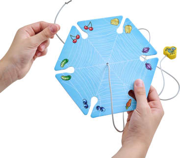 Zwei Hände halten eine sechseckige blaue Karte mit einem weißen Spinnennetzmuster, Lebensmittelillustrationen an jeder Ecke und einer durch Schlitze geführten Schnur. Ein gelber Würfel mit Bildern ist ebenfalls zu sehen.