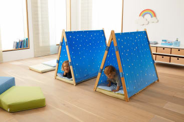 Zwei kleine Kinder spielen in kleinen blauen Stoffzelten mit weißen Sternen in einem hellen Raum mit Holzfußboden, großen Fenstern und einer an der Wand befestigten Regenbogendekoration. In der Nähe liegen Kissen auf dem Boden.