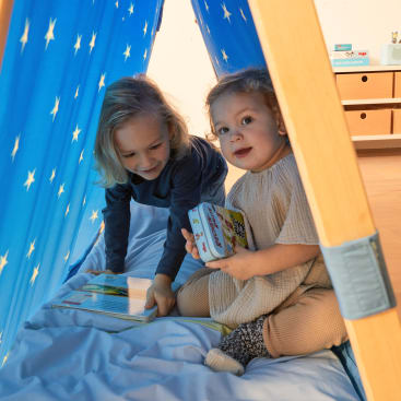Zwei kleine Kinder sitzen in einem hölzernen Spielzelt mit blauem, mit Sternenmustern bedecktem Stoff. Ein Kind hält ein Buch in der Hand, während das andere die Seiten eines aufgeschlagenen Buches auf dem Boden des Zeltes betrachtet.