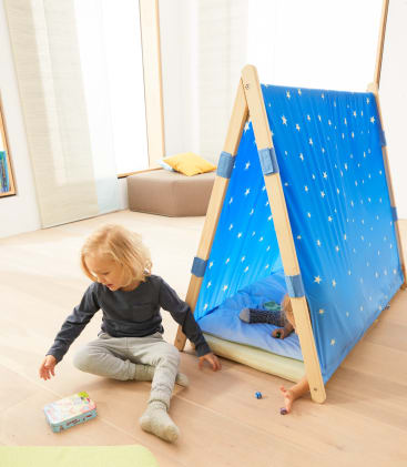 Zwei kleine Kinder spielen in einem Haus. Ein Kind sitzt in grauer Kleidung auf dem hellen Holzboden und betrachtet eine kleine Schachtel. Das andere ist meist in einem blauen, mit Sternen verzierten A-Rahmen-Spielzelt versteckt.
