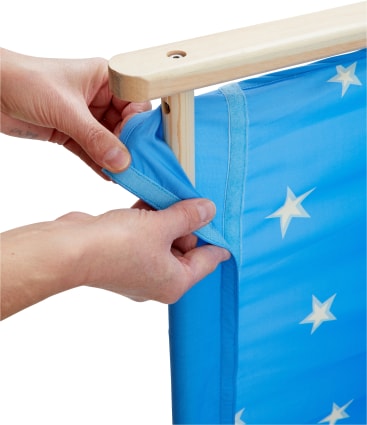 Eine Person zieht einen blauen Stoff mit weißen Sternen auf einen Holzrahmen und befestigt ihn, indem sie den Stoff über die Kante des Rahmens wickelt.