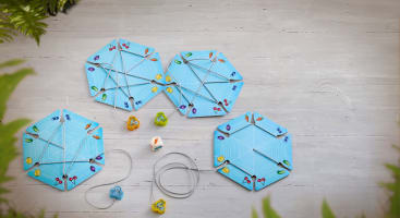 Drei blaue sechseckige Spielbretter mit bunten Fußabdrücken sind durch graue Schnüre miteinander verbunden. Vier kleine Tierfiguren und zwei Würfel stehen neben den Brettern auf einer hellen Holzfläche, die von grünen Blättern eingerahmt wird.