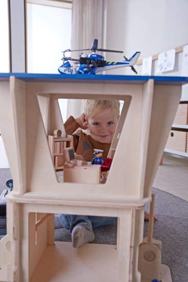 Ein kleiner Junge sitzt auf dem Boden hinter einem Holzspielzeug und schaut durch eine Öffnung. Ein Spielzeug-Polizeihubschrauber befindet sich oben auf der Struktur, und verschiedene kleine Spielzeuge befinden sich darin. Im Hintergrund sind Zeichnungen zu sehen.