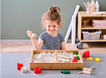 Ein kleines Kind sitzt an einem Tisch und spielt mit weißem kinetischem Sand in einem Holztablett, wobei es Holzwerkzeuge und bunte Plastikformen in Form von Gebäuden und Tieren verwendet. Im Hintergrund befinden sich Regale mit Malutensilien.