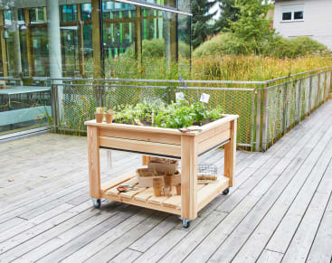 Ein hölzernes Hochbeet auf Rädern mit Grünpflanzen, Gartengeräten und Töpfen auf einer Terrasse vor einem modernen Gebäude mit Glaswänden und Grünpflanzen im Hintergrund.