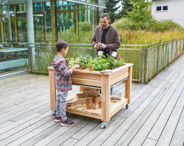 Ein Erwachsener und ein Kind stehen im Freien auf einer Holzterrasse und arbeiten mit Topfpflanzen auf einem hölzernen Gartentisch mit Rädern. Der Hintergrund zeigt ein modernes Glasgebäude und Grünflächen.