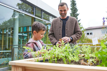 Ein Erwachsener und ein Kind stehen an einem Hochbeet im Freien und kümmern sich um verschiedene Grünpflanzen. Der Erwachsene lächelt, während das Kind die Pflanzen untersucht. Im Hintergrund sind moderne Gebäude und Bäume zu sehen.