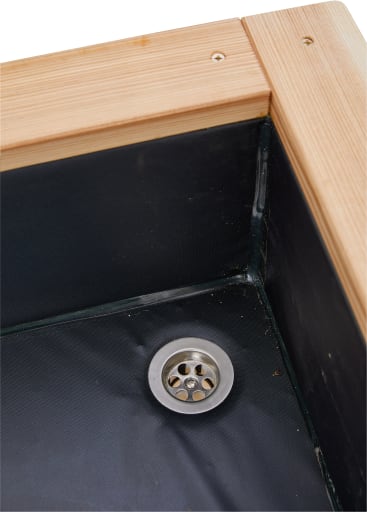 Nahaufnahme eines Metallablaufs in der Ecke einer wasserdichten Holzkonstruktion, möglicherweise eines Pflanzkastens oder einer Duschwanne, mit schwarzer Auskleidung und unbearbeiteten Holzkanten.