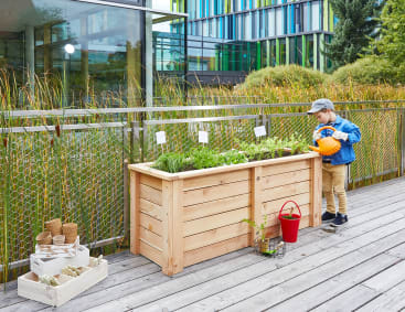 Ein kleines Kind bewässert Pflanzen in einem erhöhten Holzgartenbeet auf einer Holzterrasse neben einem Zaun. Gartengeräte und Töpfe sind in der Nähe organisiert. Im Hintergrund sind moderne Gebäude und hohe Gräser zu sehen.
