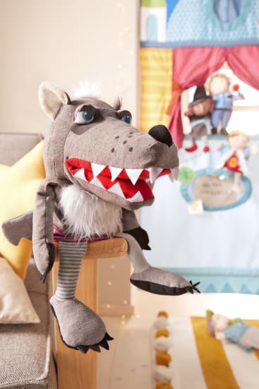 Eine große graue Wolfspuppe mit rotem Mund und weißen Zähnen sitzt auf dem Arm eines beigen Sofas. Im Hintergrund befindet sich ein buntes Stoffpuppentheater mit menschlichen Puppen und einem dekorativen Teppich auf dem Boden.