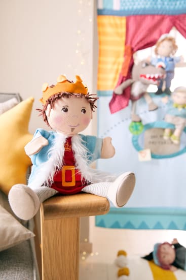 Eine Plüschpuppe, gekleidet als König mit Krone, blauem Umhang und rotem Outfit, sitzt auf einer Holzfläche. Im Hintergrund befinden sich weitere Puppen und eine bunte Stoffszene, die an der Wand hängt.
