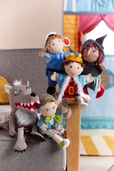 Fünf Plüschpuppen, die Märchenfiguren darstellen, darunter ein Wolf, eine Großmutter, ein Prinz, eine Hexe und ein Junge in Blau, sind auf einer grauen Couch und einer hölzernen Armlehne in einer farbenfrohen Spielzimmerumgebung angeordnet.