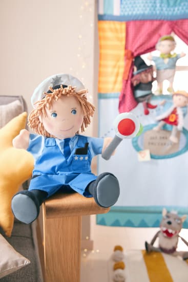 Eine als Polizist verkleidete Plüschpuppe sitzt auf der Armlehne eines Holzstuhls und hält einen Spielzeugschlagstock. Im Hintergrund ist ein buntes Puppentheater mit anderen Plüschpuppen und Spielzeug zu sehen.