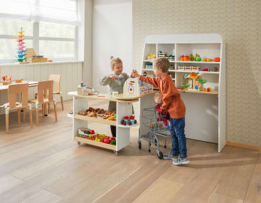 Zwei kleine Kinder spielen in einem vorgetäuschten Lebensmittelladen. Eines steht hinter der Theke mit Spielzeuglebensmitteln, während das andere einen Einkaufswagen mit einer Puppe schiebt. Der Hintergrund zeigt Regale mit Spielobst und -gemüse.