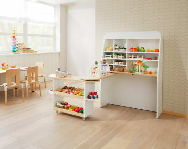 Ein Kinderspielzimmer mit einem hölzernen Spielmarktstand, der mit Spielzeugobst, -gemüse und -lebensmitteln bestückt ist. Kleine Holztische und Stühle stehen im Hintergrund neben großen Fenstern und einer gemusterten Wand.