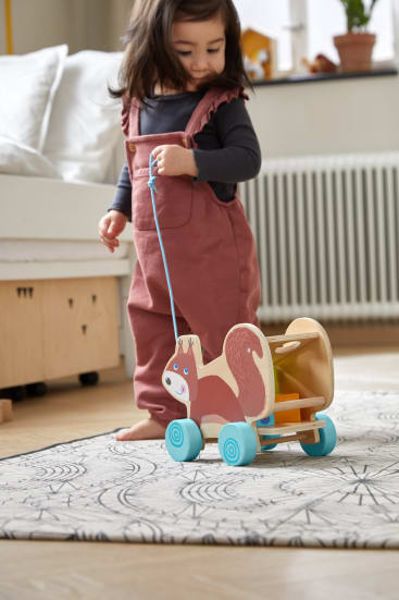 Ein kleines Kind in einem rosa Overall zieht ein Holzspielzeug in Form eines Eichhörnchens auf Rädern über einen gemusterten Teppich in einem hellen Zimmer mit einem Bett und einem Heizkörper im Hintergrund.