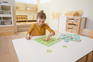 Ein junges Mädchen sitzt an einem weißen Tisch in einem Klassenzimmer und spielt ein pädagogisches Brettspiel mit illustrierten Karten. Im Hintergrund sind Holzregale und Schränke zu sehen.