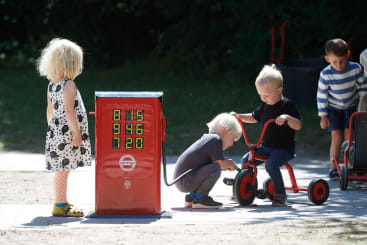 Drei kleine Kinder spielen im Freien mit einer roten Spielzeug-Tanksäule und Dreirädern. Ein Kind hält die Zapfpistole, ein anderes hockt, und ein drittes steht in einem weißen Kleid mit schwarzen Flecken in der Nähe. Zwei weitere Kinder sind im Hintergrund zu sehen.
