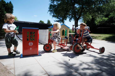 Drei kleine Kinder spielen im Freien mit Spielzeugdreirädern neben einer Spieltanksäule auf einer gepflasterten Fläche. Im Hintergrund sind ein kleines Spielhaus und Bäume zu sehen.