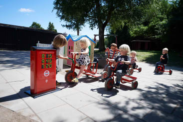 Mehrere kleine Kinder fahren mit Dreirädern und Tretautos im Freien auf einer gepflasterten Fläche. Ein Kind drückt auf Knöpfe an einer roten Spiel-Tankstelle. Im Hintergrund sind Bäume, Gras und ein Spielhaus zu sehen.