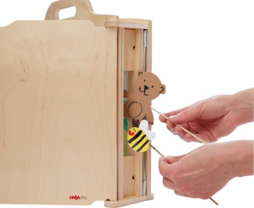 Ein Paar Hände hält mit Holzstäben eine Bären- und eine Bienenpuppe hinter einer hölzernen Erzählkiste mit seitlicher Teilöffnung. Die Schachtel hat einen Griff und das HABA Pro-Logo auf ihr.