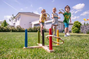 Drei Kinder stehen an einem sonnigen Tag vor einem modernen Haus auf einer Wiese und werfen Seilringe in Richtung eines hölzernen Ringwurfspiels mit farbigen Pflöcken.