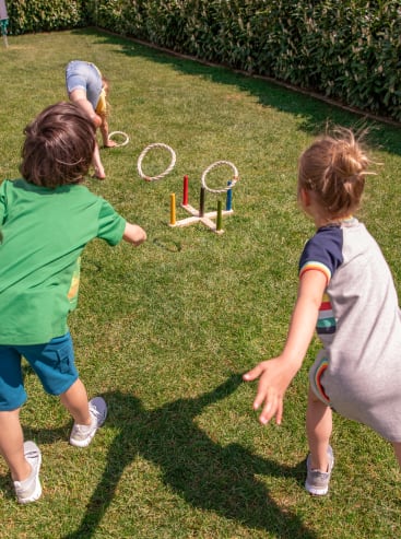 Drei Kinder spielen ein Ringwurfspiel auf einer Wiese. Zwei Kinder im Vordergrund werfen Ringe in Richtung eines hölzernen Ringwurfsets, während sich ein Kind im Hintergrund bückt, um einen Ring aufzuheben.