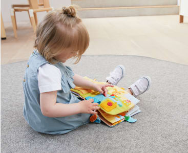 Ein kleines Kind sitzt im Schneidersitz auf einem grauen Teppich, trägt ein blaues Kleid und weiße Schuhe, blickt nach unten und spielt mit einem bunten Activity-Buch aus Stoff.